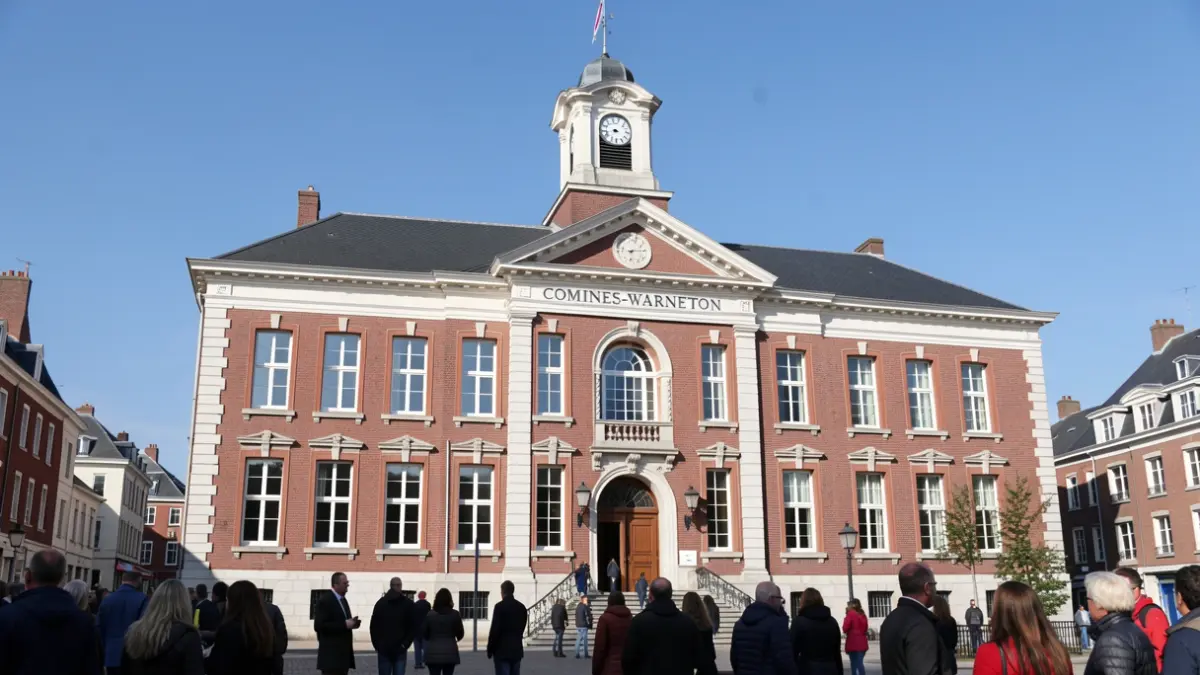 Facade of the Comines-Warneton town hall, a red brick building with a white stone trim and a clock tower, under a clear blue sky.